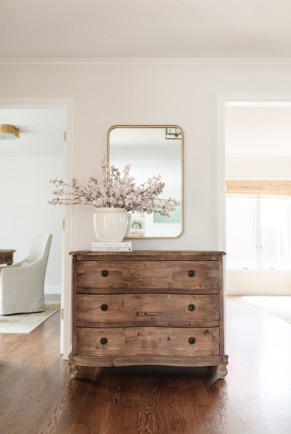 A wooden chest with a brass mirror above in the entryway of a light filled home
