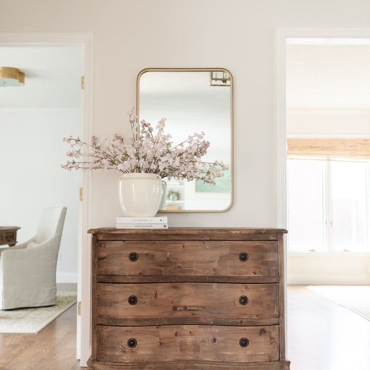 A wooden chest with a brass mirror above in the entryway of a light filled home