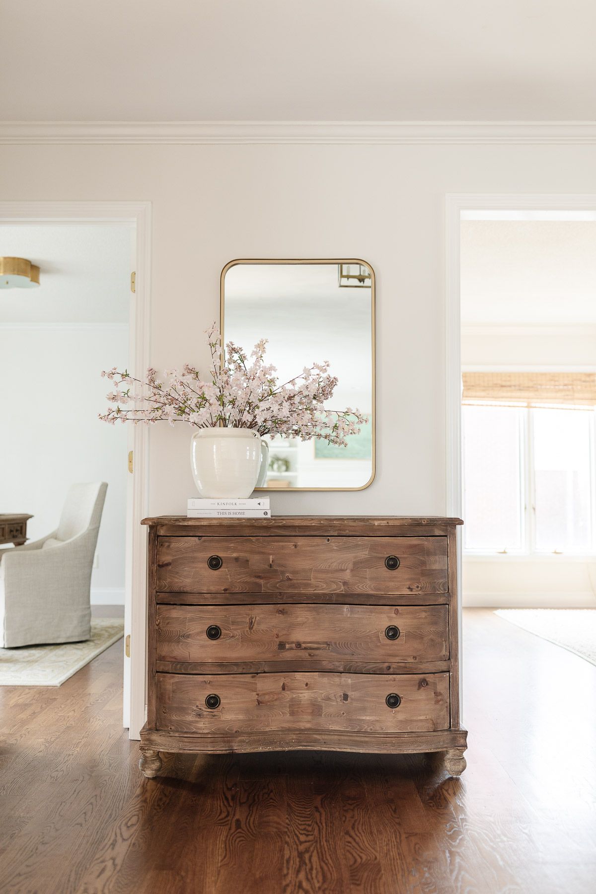 A wooden chest with a brass mirror above in the entryway of a light filled home