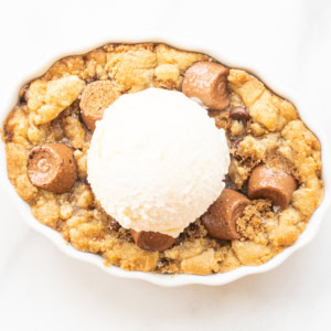 A chocolate chip cobbler in an oval dish topped with a scoop of vanilla ice cream, chocolate candy pieces, and a crumbly cookie mixture.