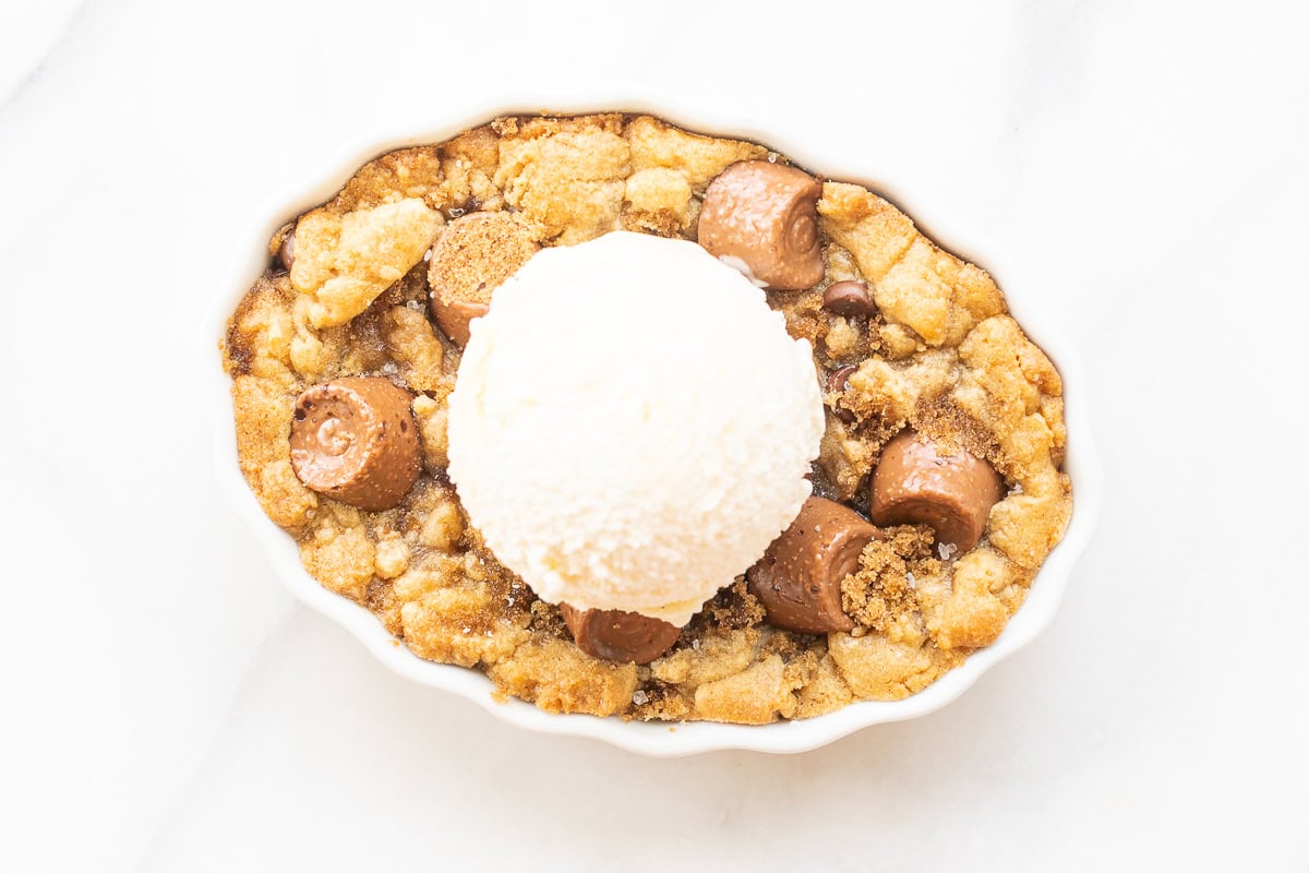 A dessert dish featuring a chocolate chip cookie cobbler base topped with chocolate pieces and a single scoop of vanilla ice cream on a white background.