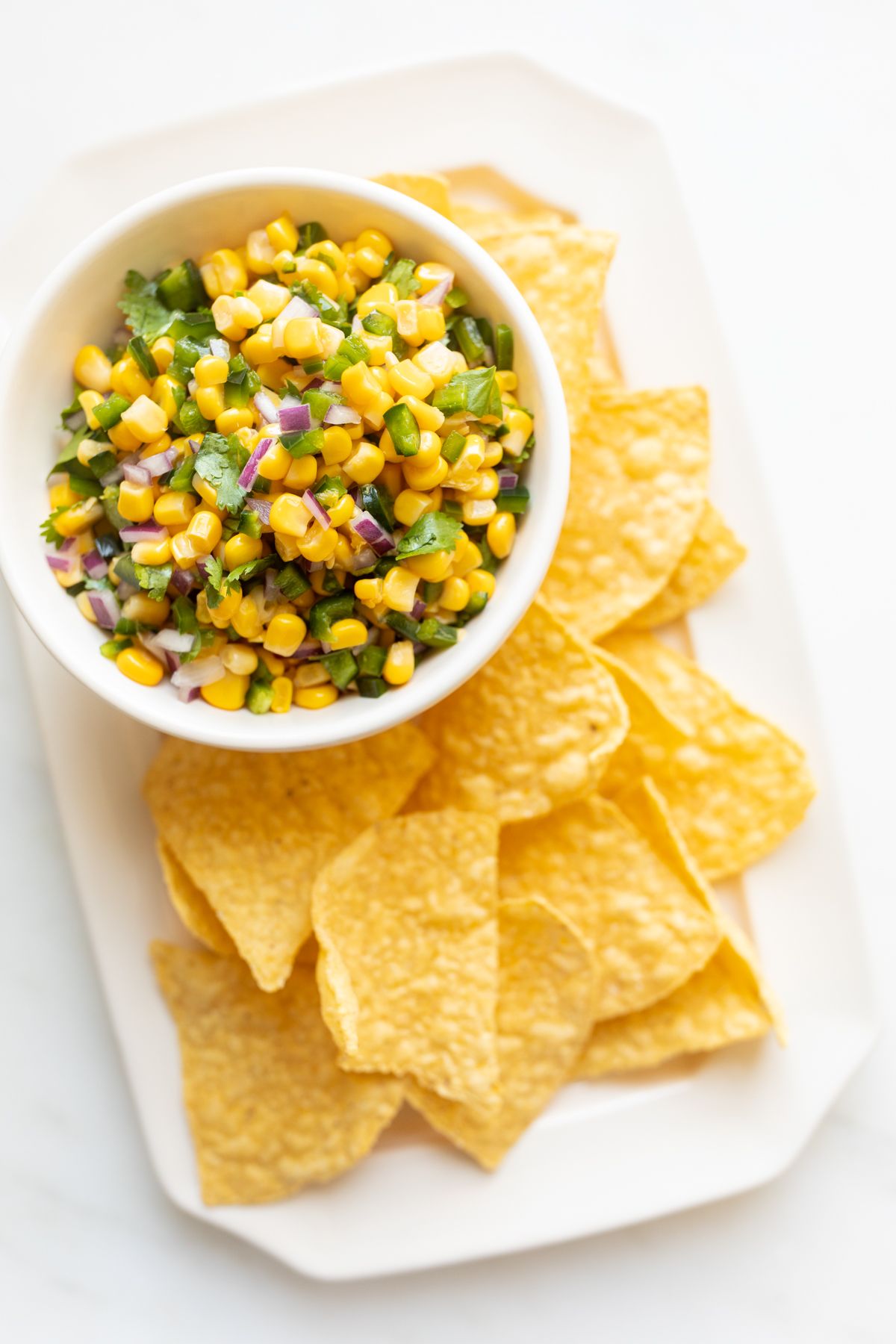 A chipotle corn salsa recipe in a white bowl, placed on a white platter full of corn tortilla chips