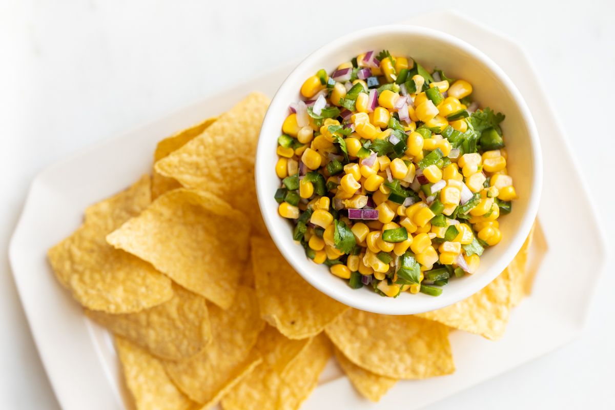 A chipotle corn salsa recipe in a white bowl, placed on a white platter full of corn tortilla chips
