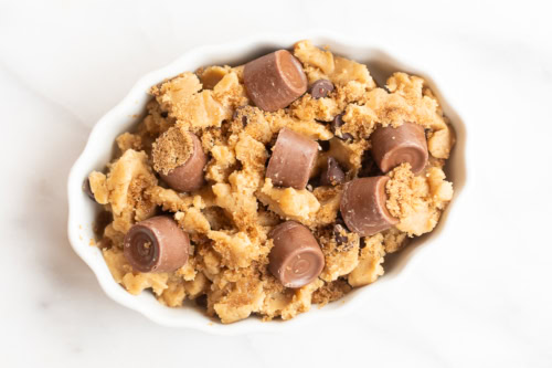 A white bowl filled with cookie dough, chocolate chips, and chocolate candy pieces nestled into the dough sits on a white surface, evoking the delightful essence of a chocolate chip cobbler.