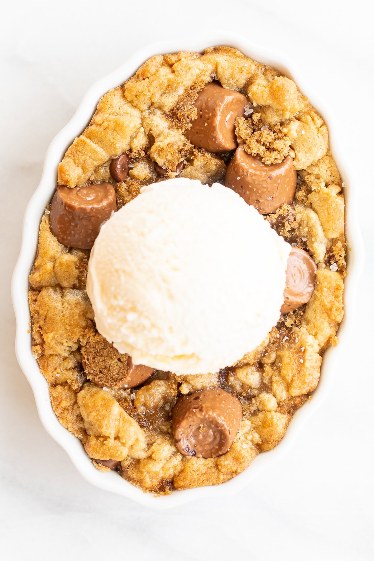 A round caramel cobbler with a crumbly topping, pieces of caramel chocolates, and a scoop of vanilla ice cream on top in a white dish.