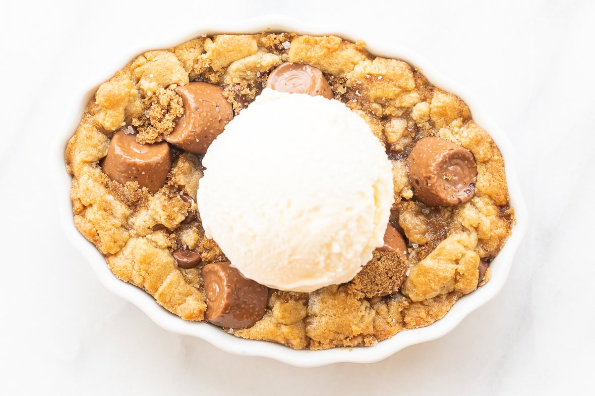 A white dish contains a caramel cobbler with a crumbly texture, dotted with chocolate pieces, and crowned by a scoop of vanilla ice cream on top, all placed on a pristine white surface.