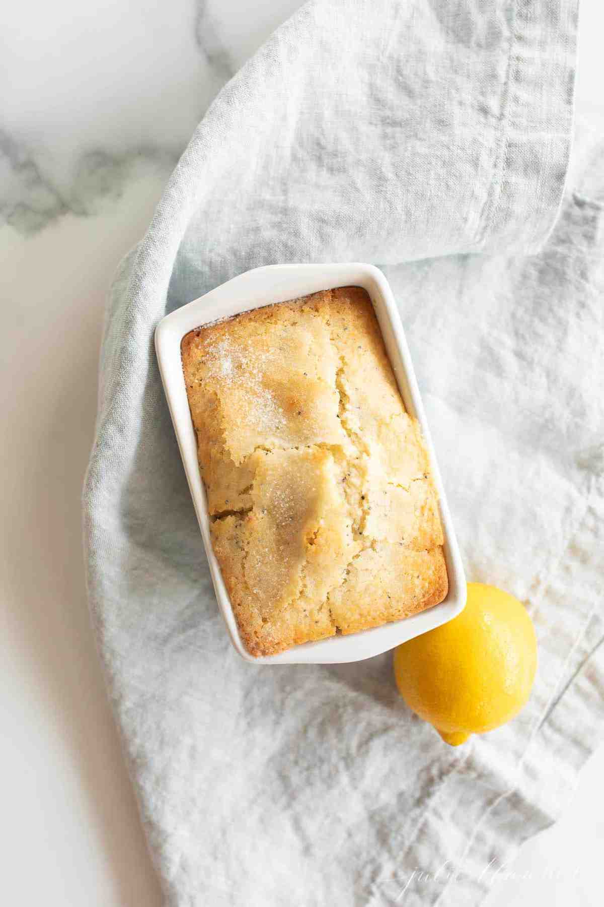 A blue linen background with a small loaf of lemon poppy seed bread recipe, lemon to the side.