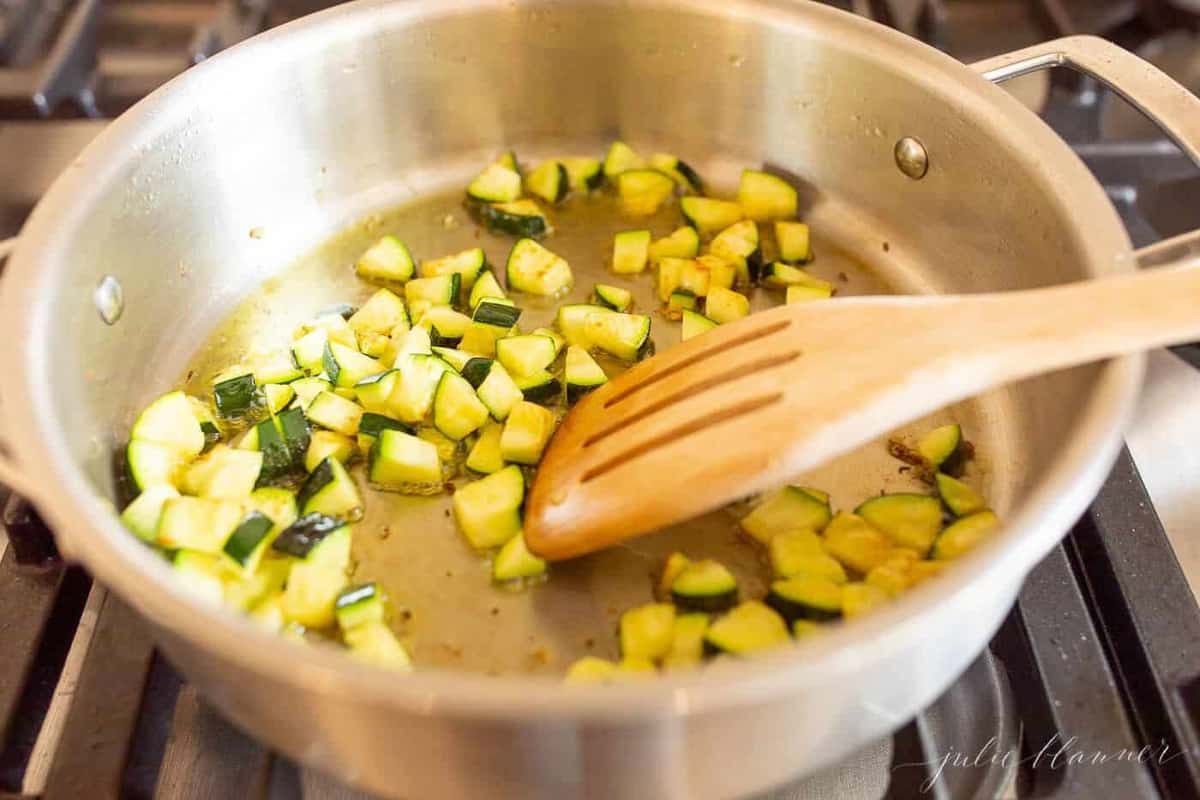 A silver stockpot, wooden spoon sauteeing squash for a pasta salad recipe.