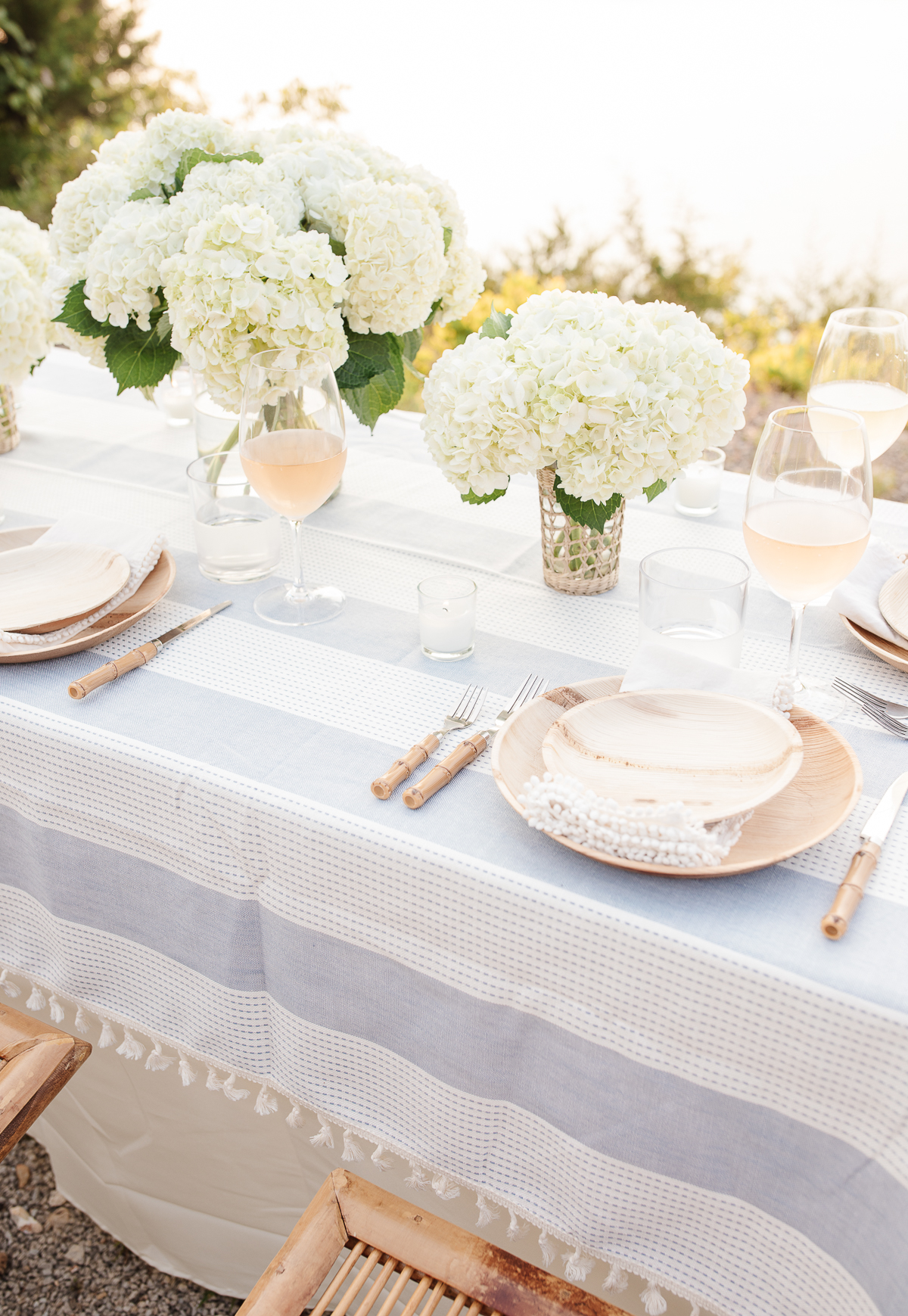 Elegant outdoor dining setup with a blue lace tablecloth, white hydrangea centerpieces, and bamboo chairs.