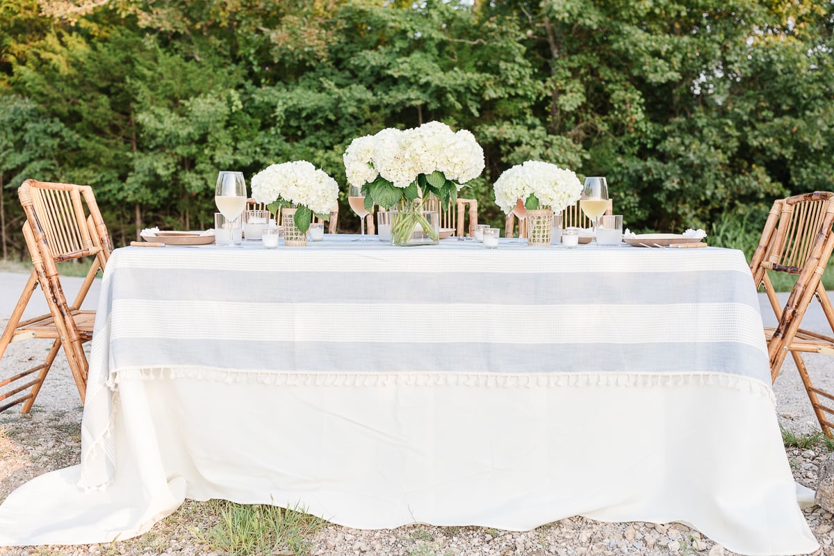 Elegant outdoor dining setup with a white tablecloth, wooden chairs, and hydrangea centerpieces on a gravel surface, surrounded by greenery.
