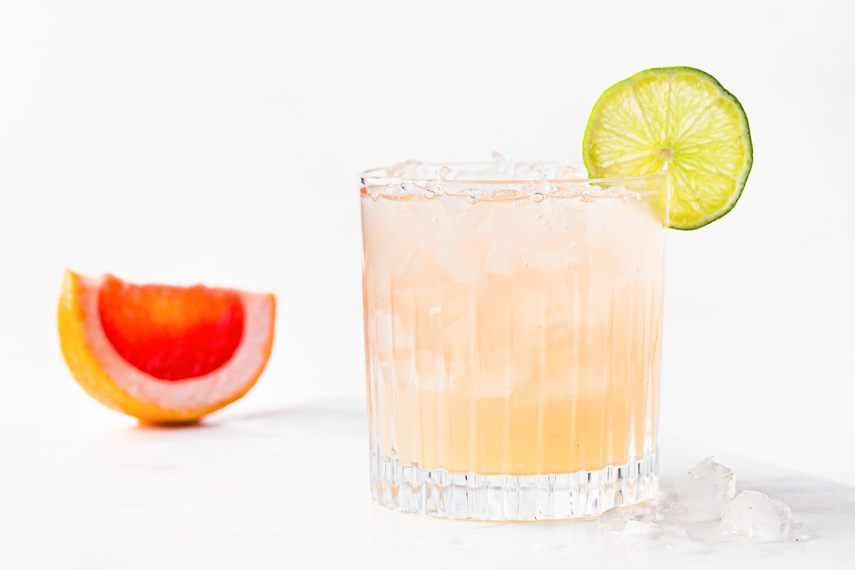 A paloma cocktail with a lime garnish and a slice off grapefruit in the background