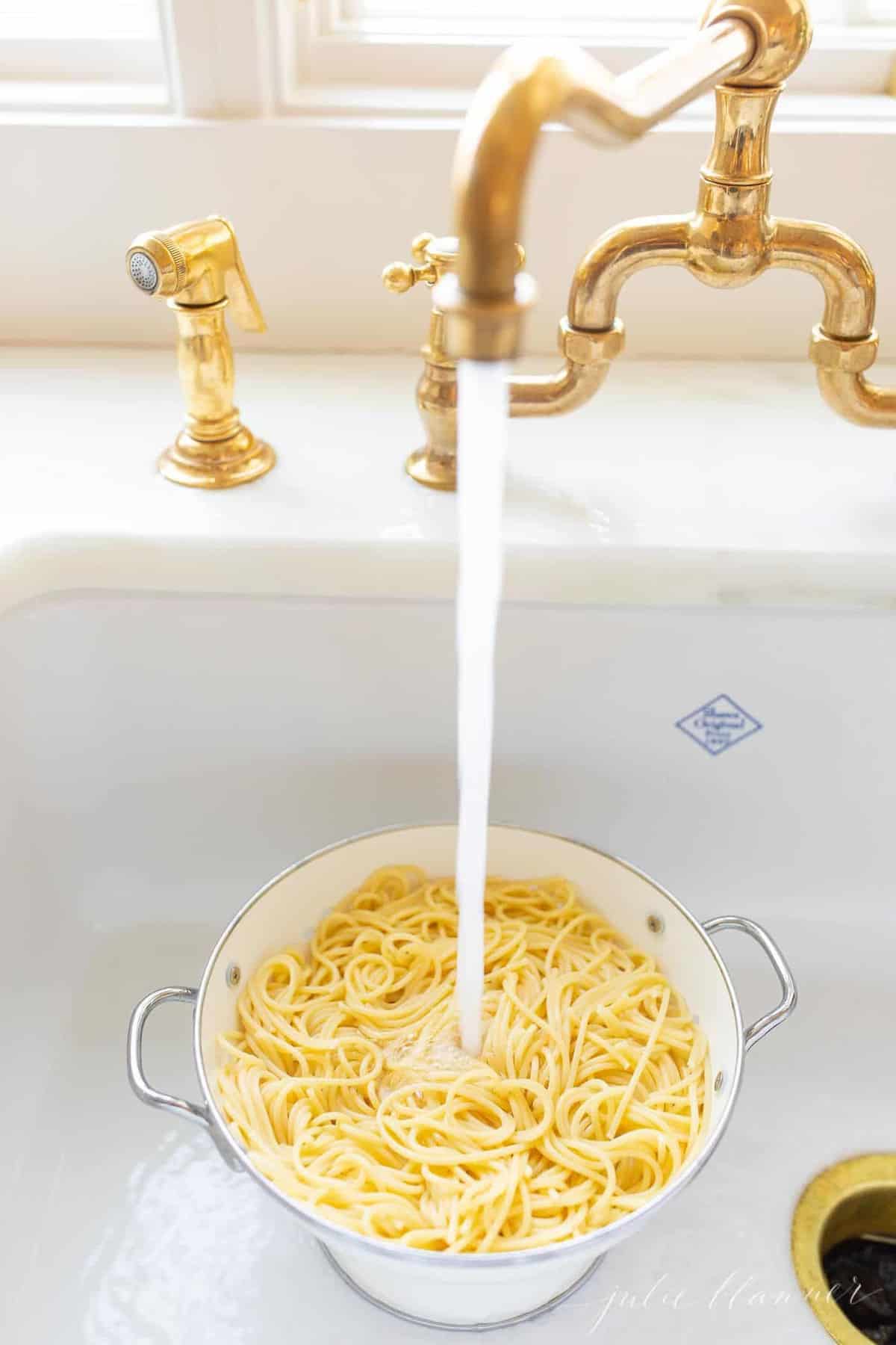 A white sink with water running into a strainer filled with cooked spaghetti.