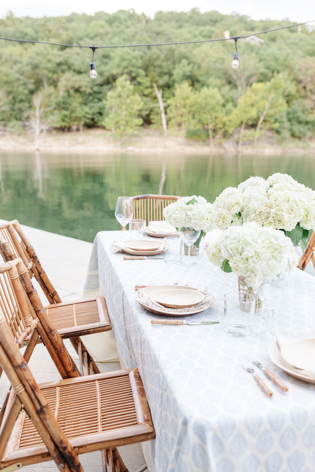 Elegant outdoor dining setup by a tranquil lake featuring a table with blue linens, wooden chairs, hydrangea centerpieces, and hanging string lights.