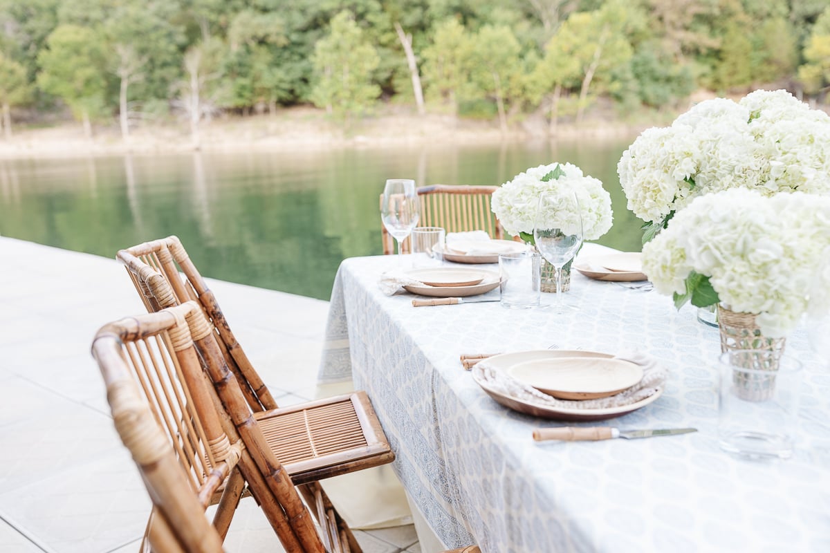 Elegant outdoor dining setup by a tranquil lake, featuring a table with fine china, glasses, and lush hydrangea centerpieces.