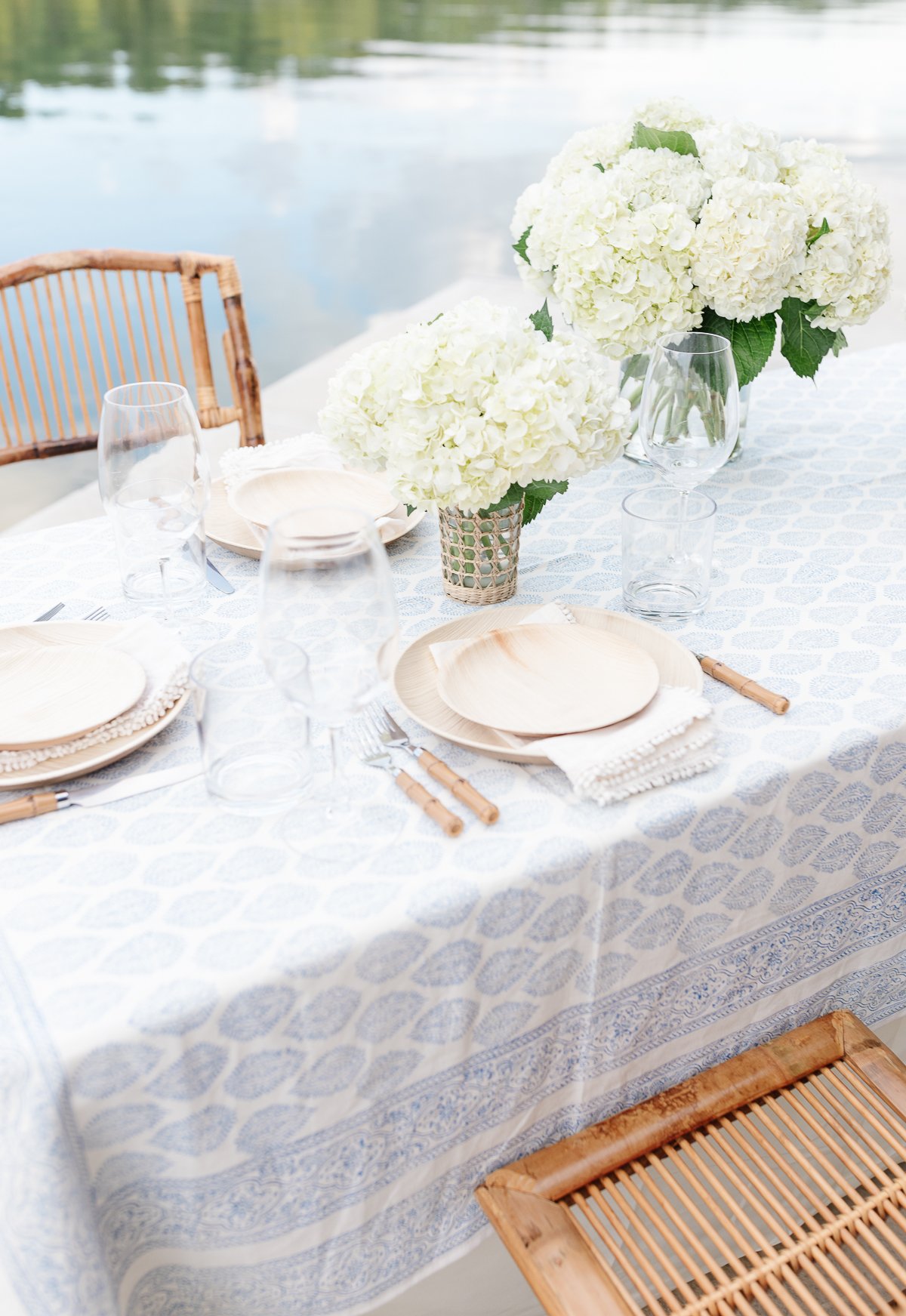 Elegant outdoor dining setup with a blue lace tablecloth, white hydrangea centerpieces, and bamboo chairs, showcasing a serene and stylish table arrangement.