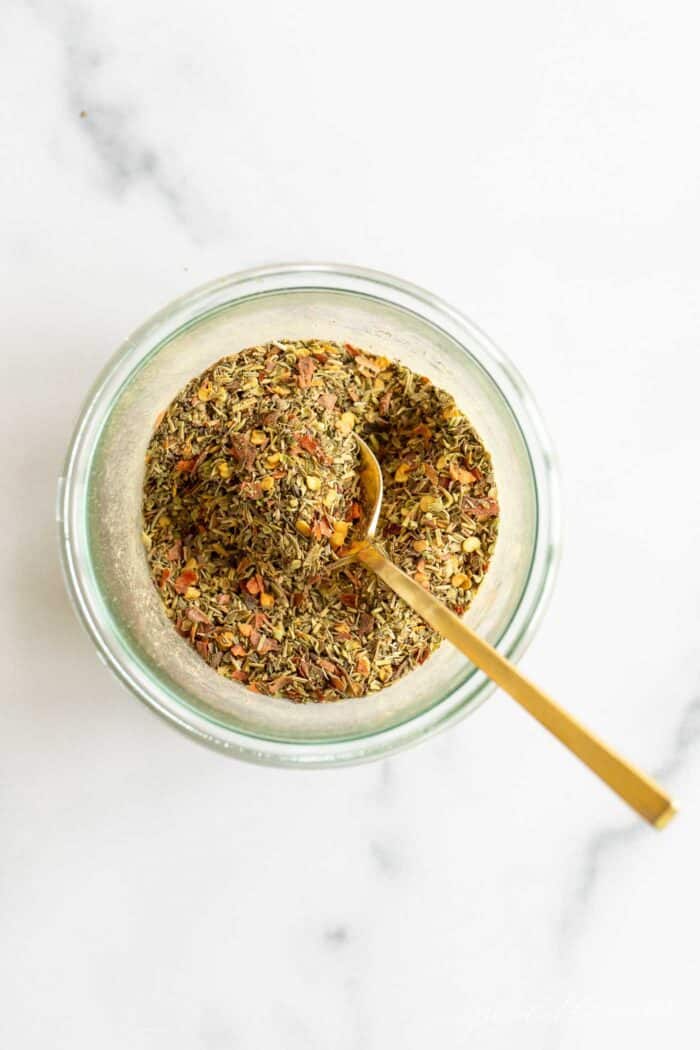 A clear glass jar of italian seasoning blend, gold spoon sticking out.