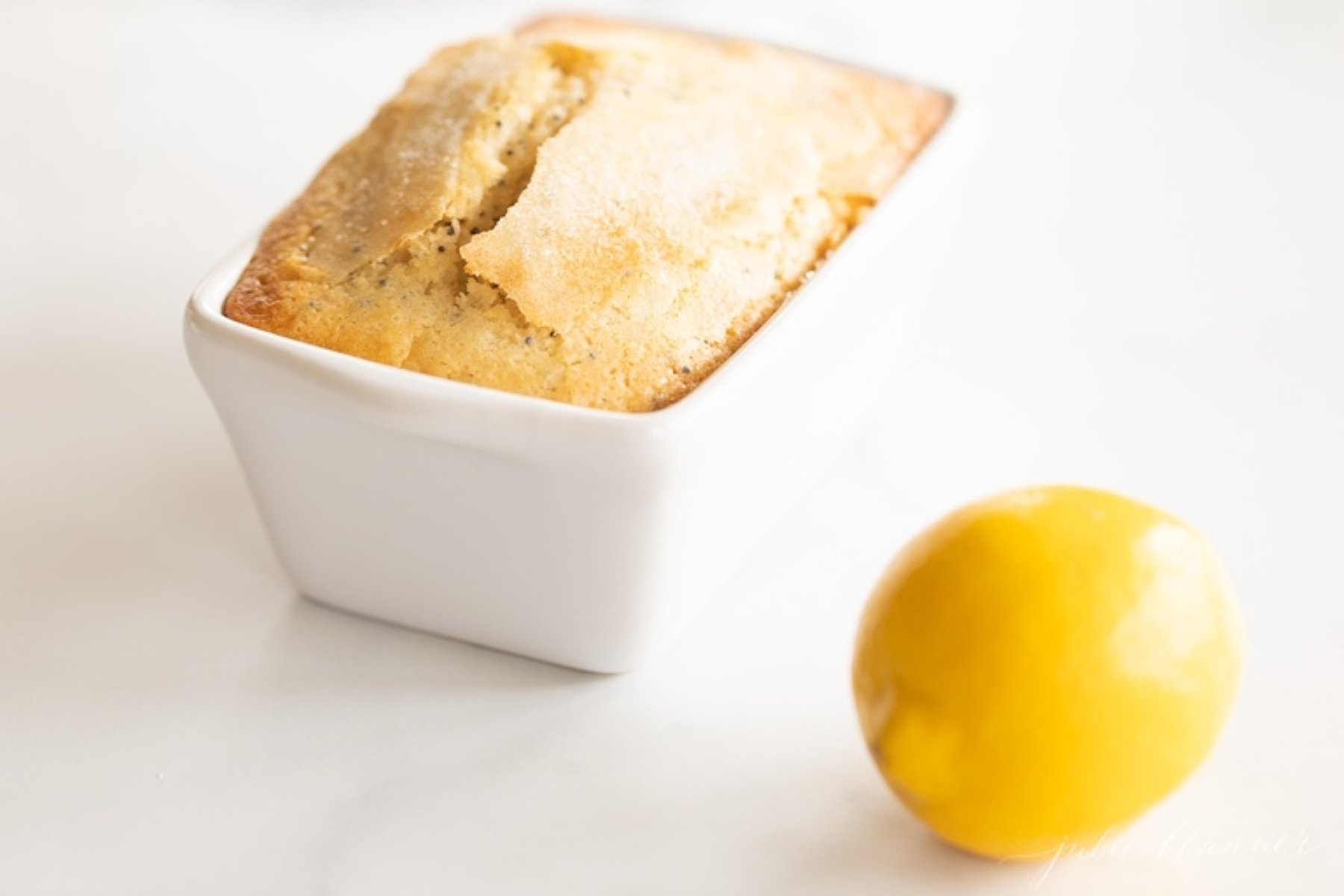 A small loaf of lemon poppy seed bread in a white ceramic loaf pan, with a lemon placed beside it. 