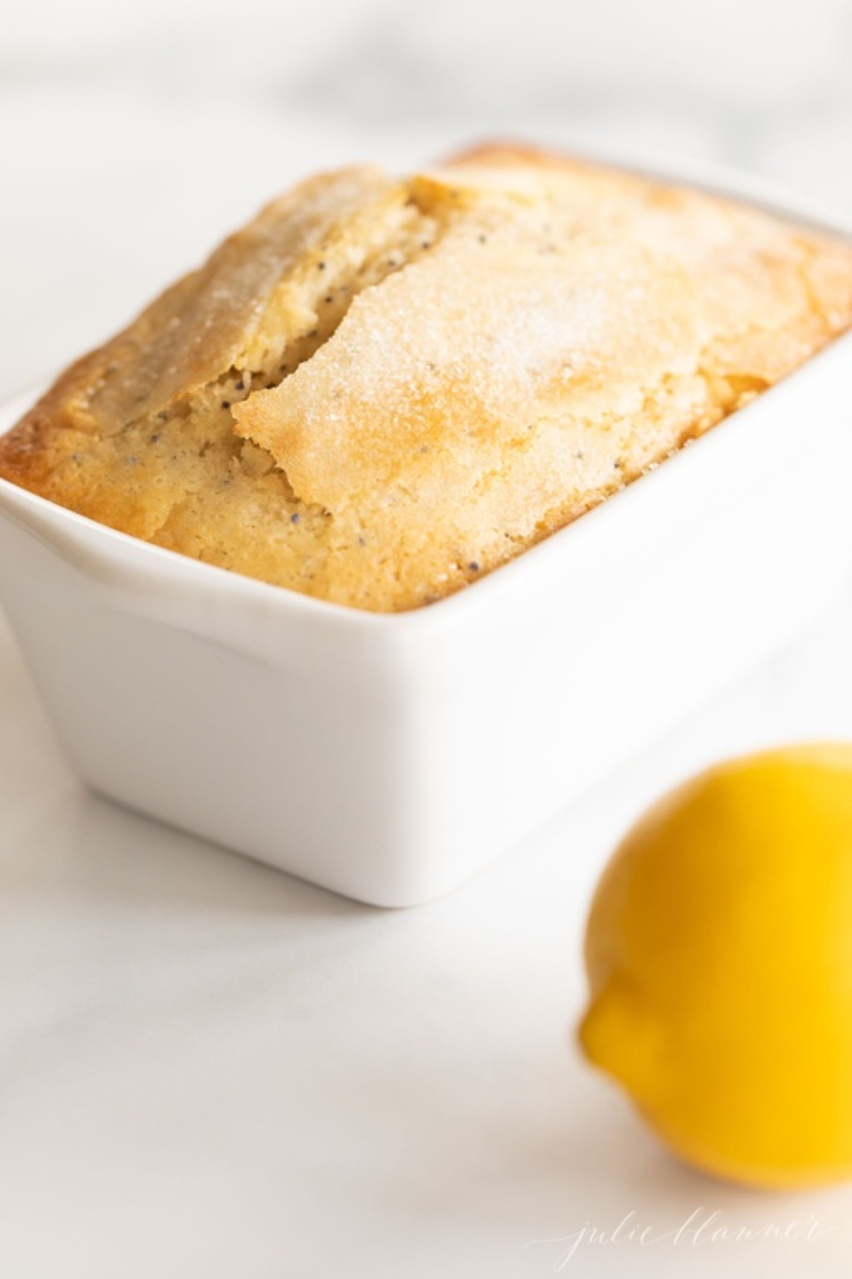 A small loaf of lemon poppy seed bread in a white ceramic loaf pan, with a lemon placed beside it.