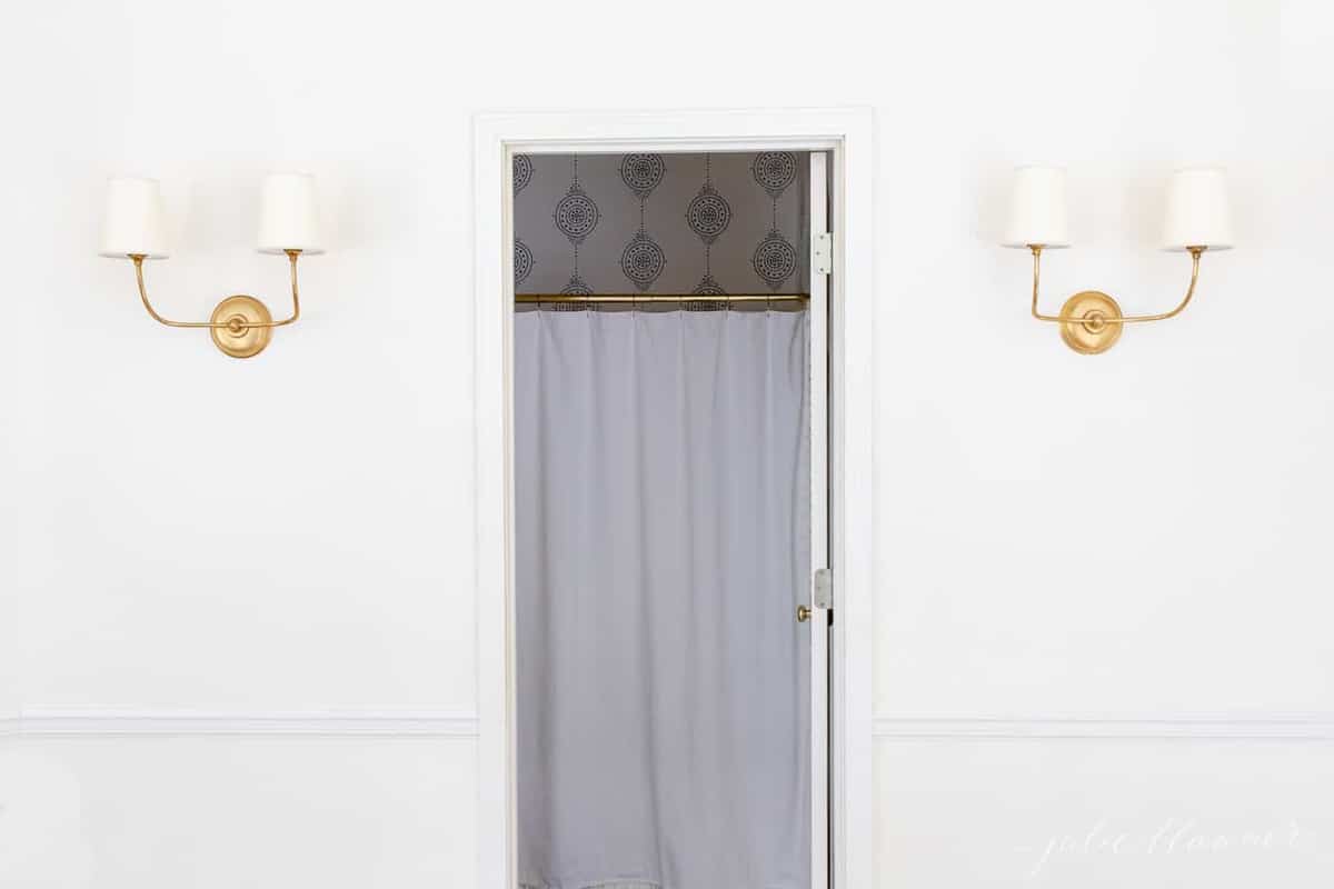 A white painted hallway with two gold wall sconces, door open into bathroom in between.