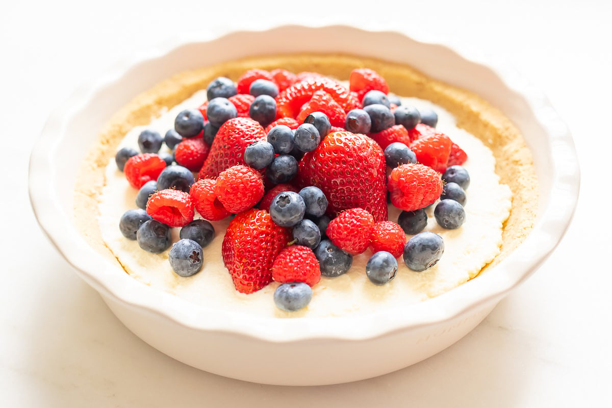 A no bake lemon pie with a creamy filling topped with fresh strawberries, raspberries, and blueberries in a white pie dish.