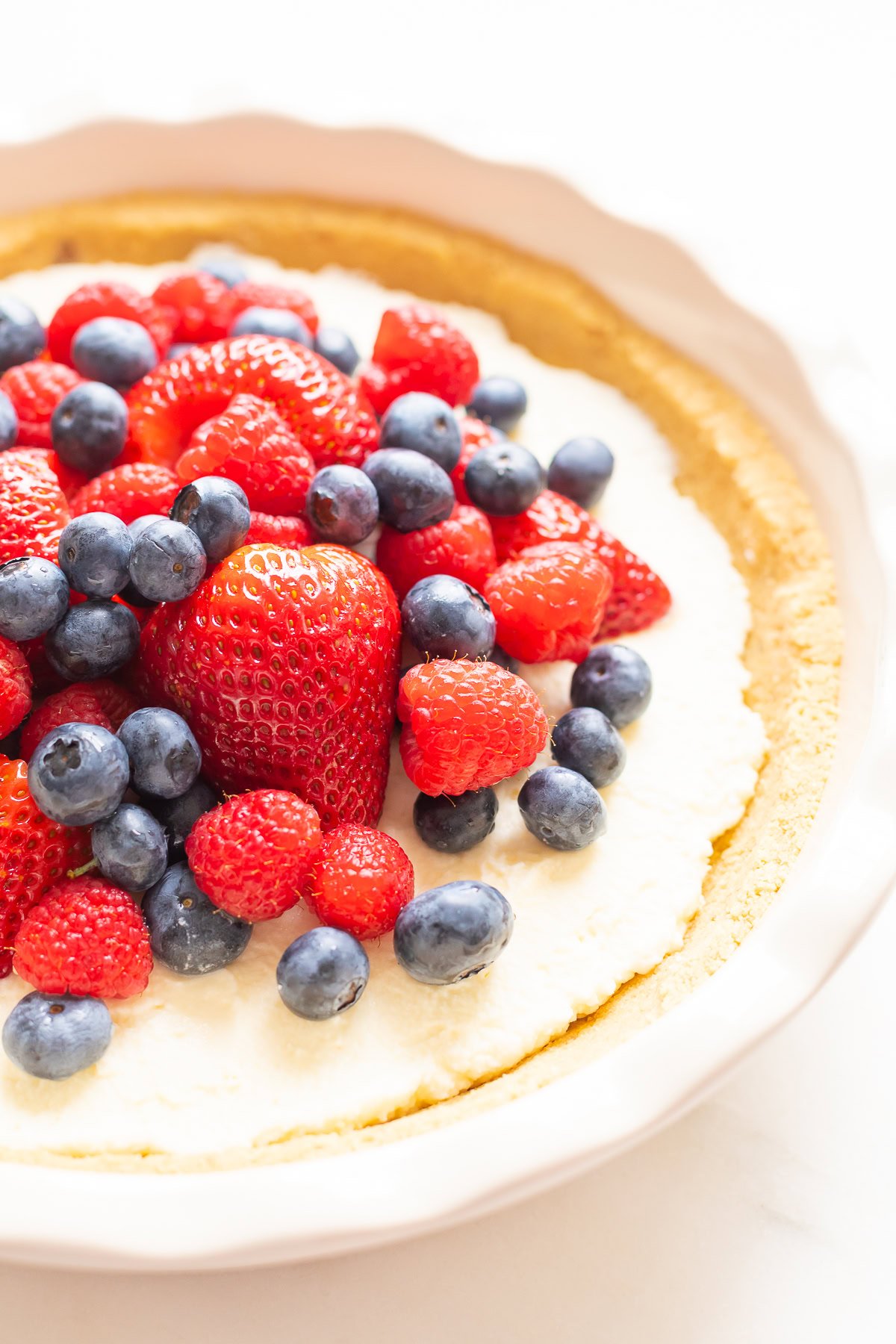 A close-up of a no bake lemon pie cheesecake topped with fresh strawberries, raspberries, and blueberries in a pie dish. The cheesecake has a graham cracker crust.