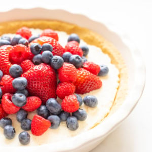 A no-bake lemon pie filled with a creamy base and topped with fresh strawberries, raspberries, and blueberries is displayed in a white ceramic dish.