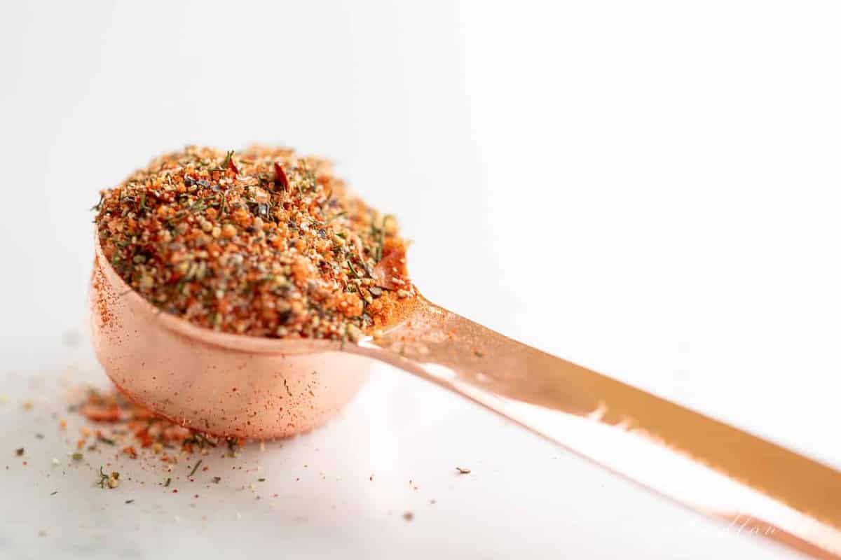 A white background with a copper measuring spoon filled with steak seasoning blend on top.