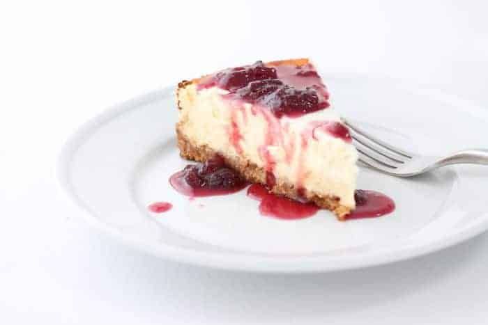 A white background and white place with a slice of cheesecake drizzled with a strawberry topping for cheesecake.