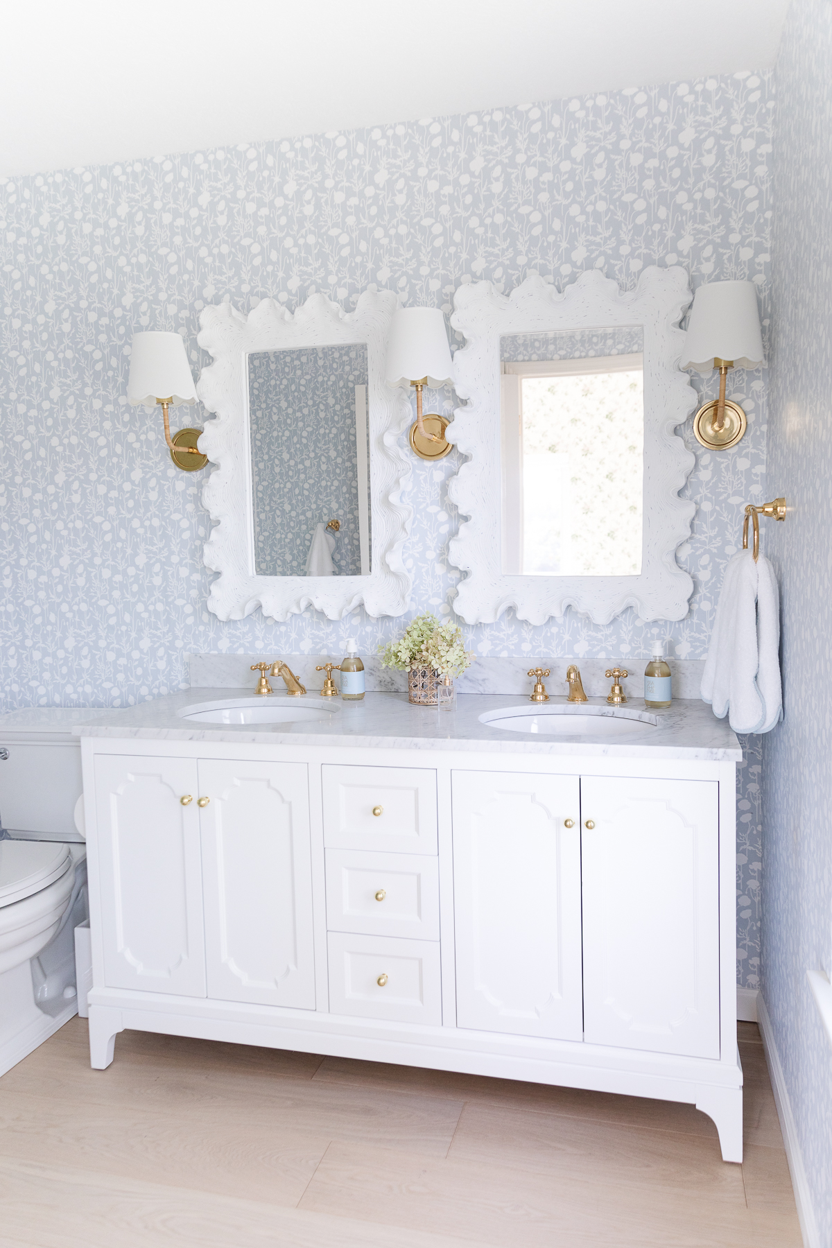 A wallpapered bathroom with a white vanity and scalloped mirrors