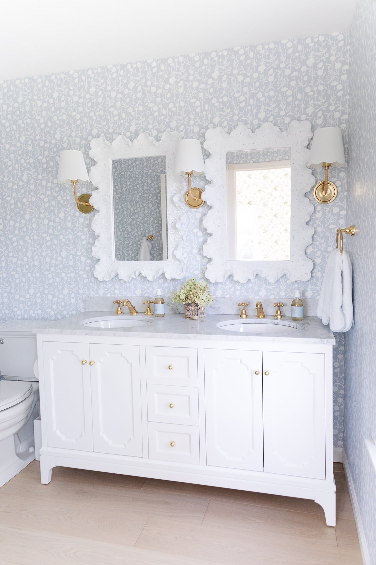 A wallpapered bathroom with a white vanity and scalloped mirrors