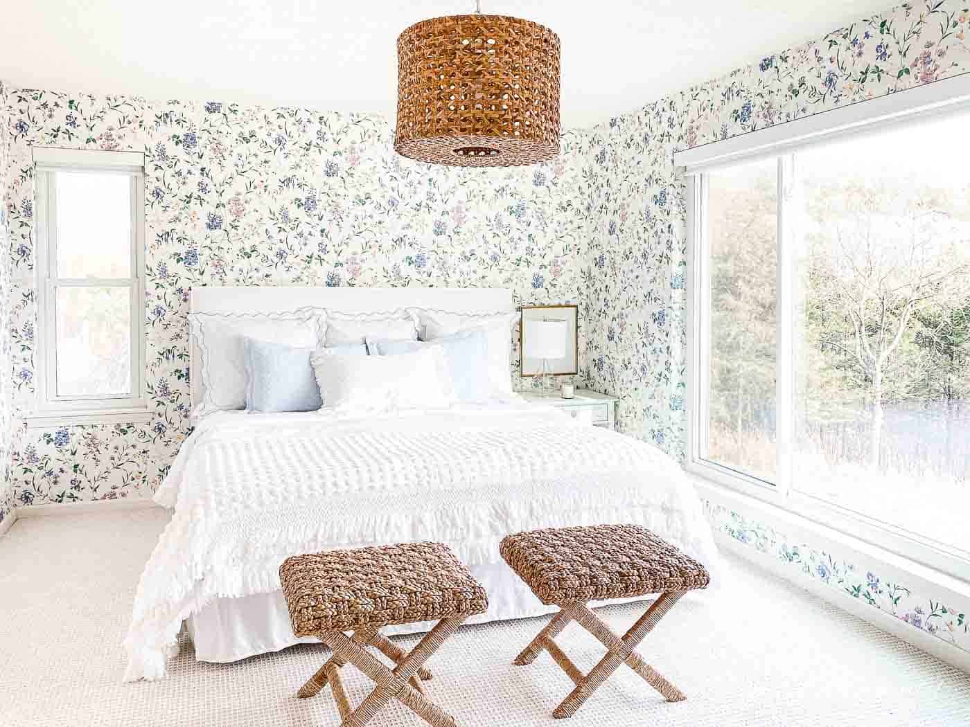 wallpapered bedroom with white slipcovered headboard, white bedding and rattan stools