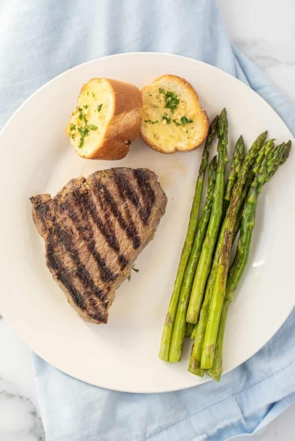 Marinated steak on a white plate with sliced bread and asparagus.