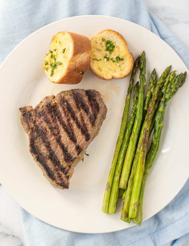 Marinated steak on a white plate with sliced bread and asparagus.