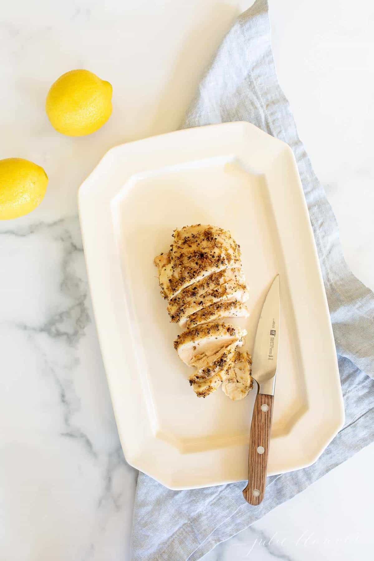 A white platter with a lemon pepper baked chicken breast sliced, knife to the side.