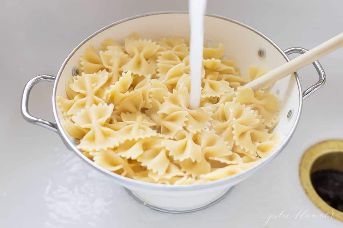 A white colander with water coming in, rinsing cooked pasta for a lesson in how to make pasta salad.