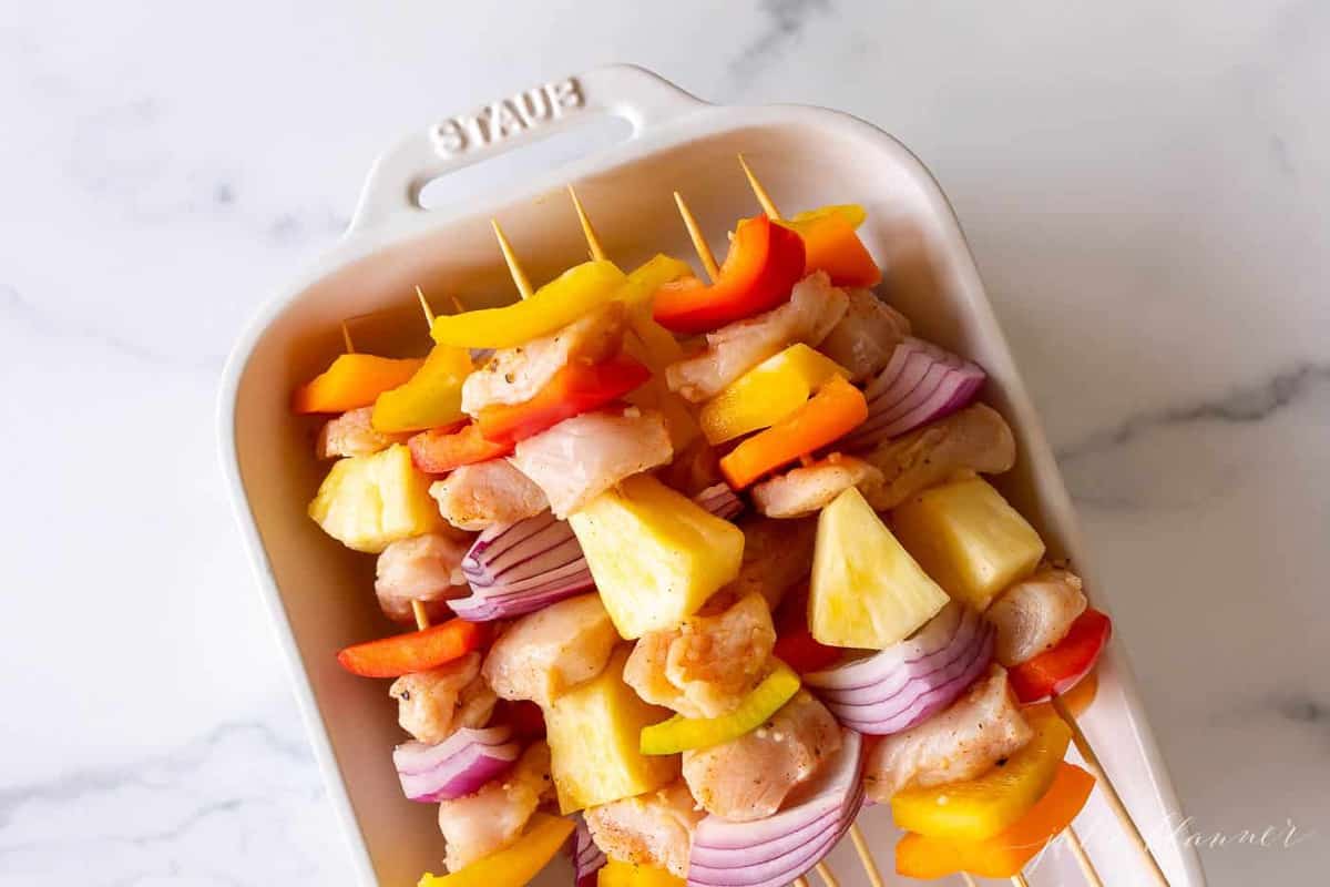 A white baking dish filled with bamboo chicken skewers, on a marble surface.