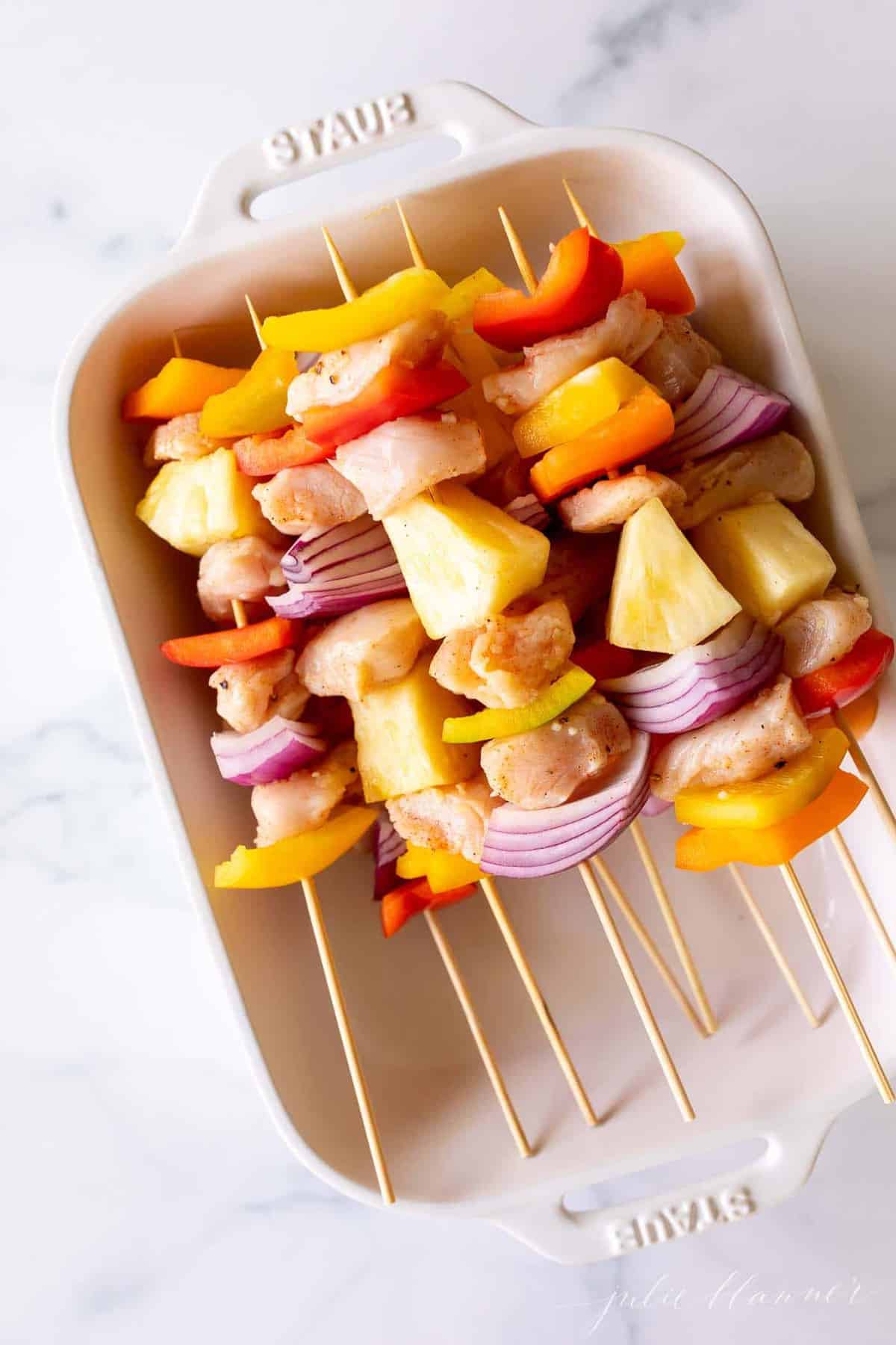 A white baking dish filled with bamboo chicken skewers, on a marble surface.