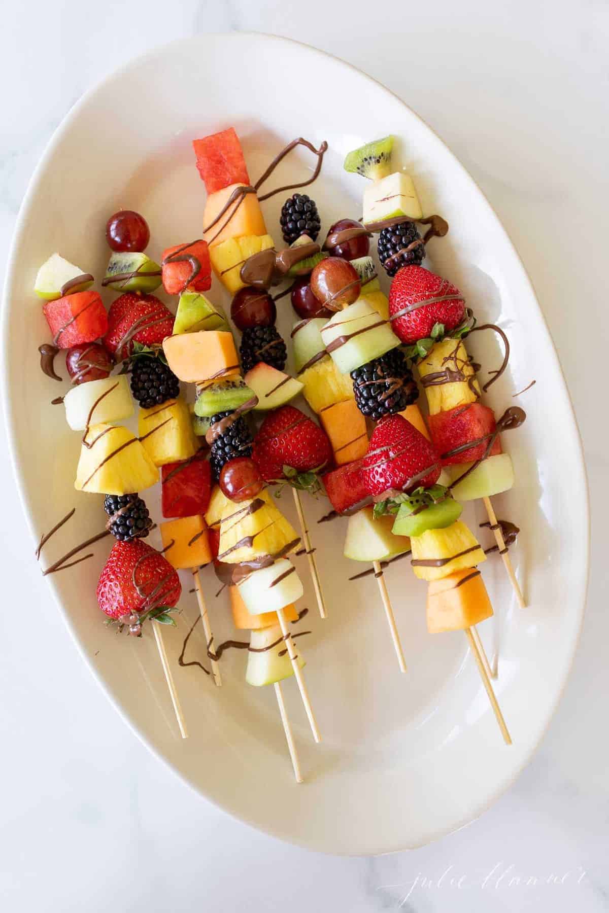 Colorful fruit skewers on a white oval platter, covered in a drizzle of chocolate.