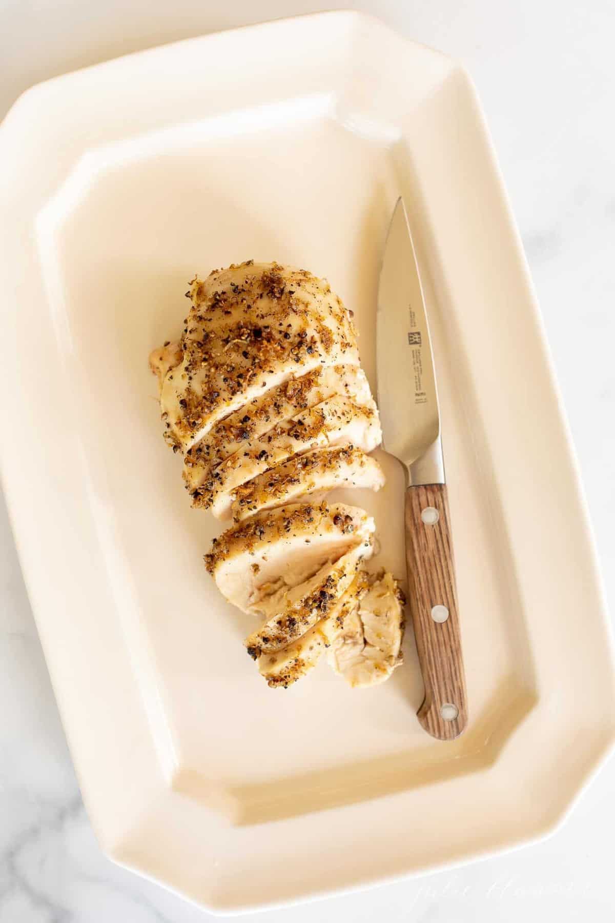 A white platter with a lemon pepper baked chicken breast sliced, knife to the side.