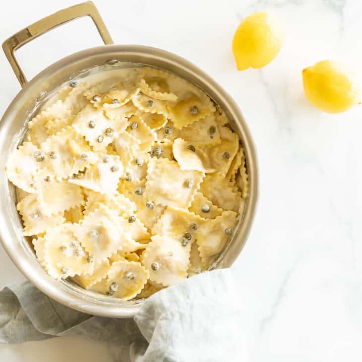 A pot full of white wine lemon caper sauce covered pasta, lemons and linen napkins surrounding it.