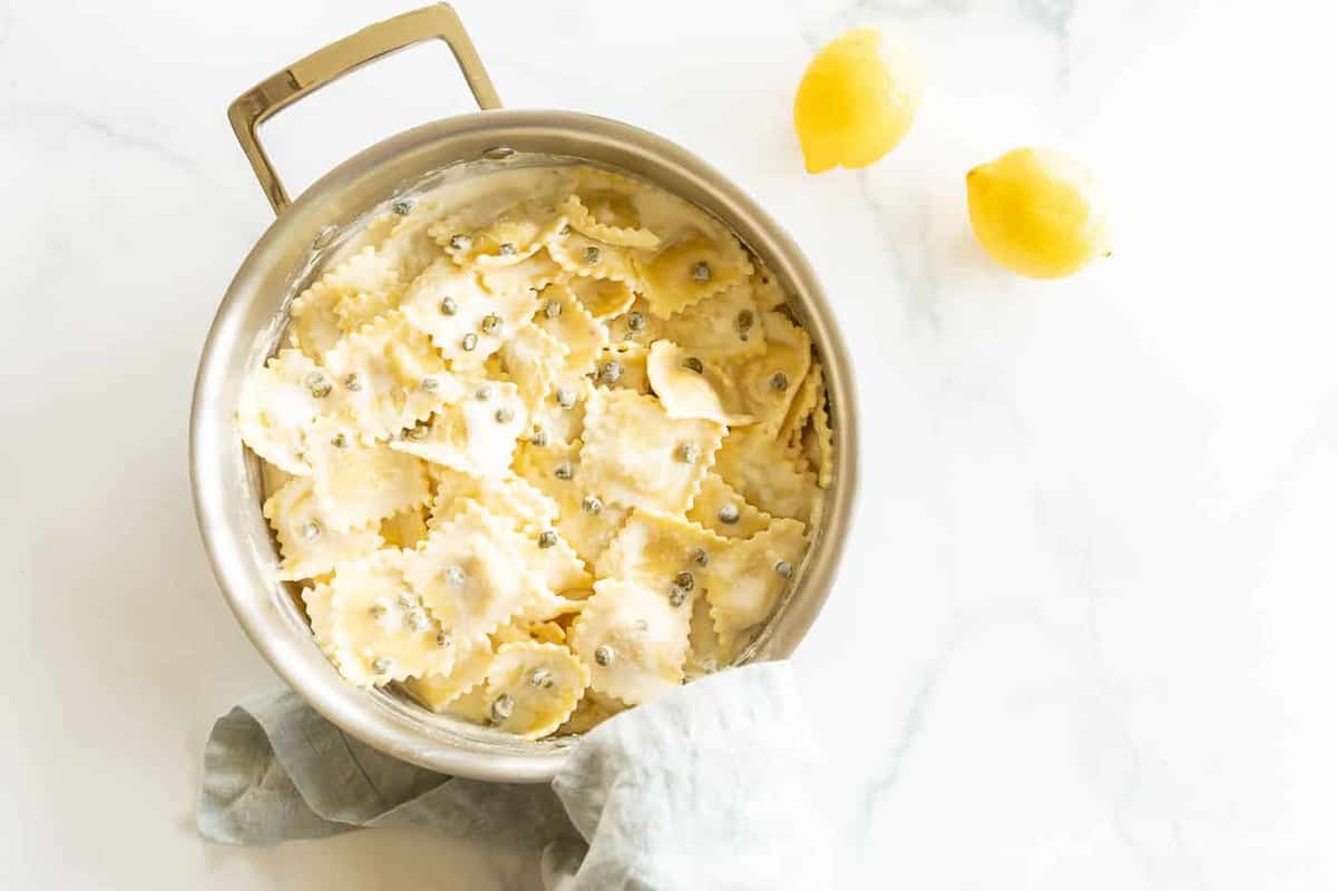 A pot full of white wine lemon caper sauce covered pasta, lemons and linen napkins surrounding it. 