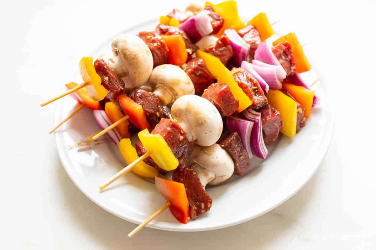 A platter full of skewers with veggies and raw steak for kabobs.