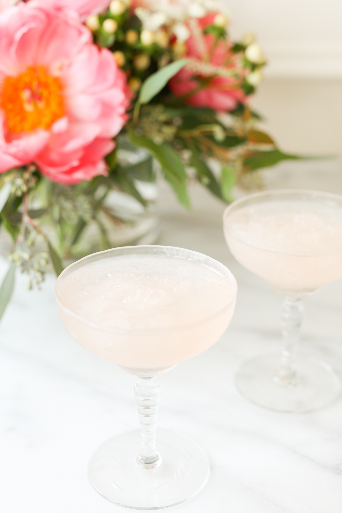 Two pink frosé cocktails in front of a bouquet of flowers.