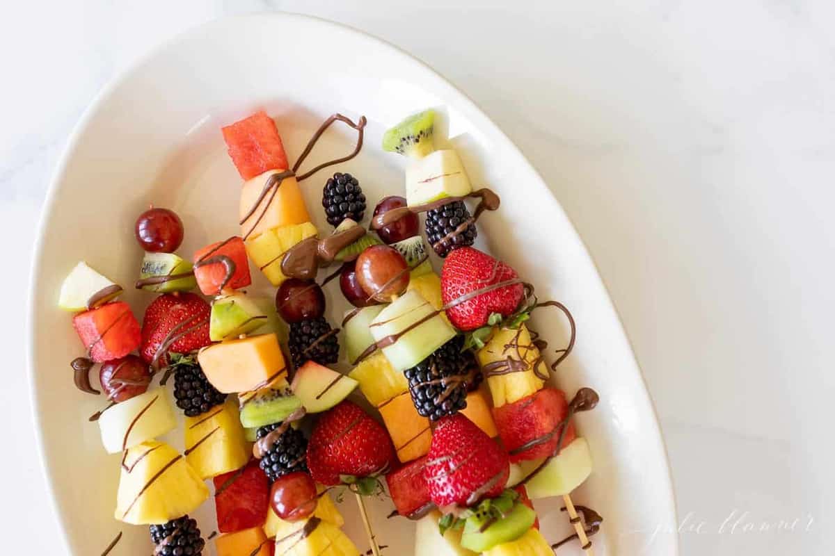 Colorful fruit skewers on a white oval platter, covered in a drizzle of chocolate.