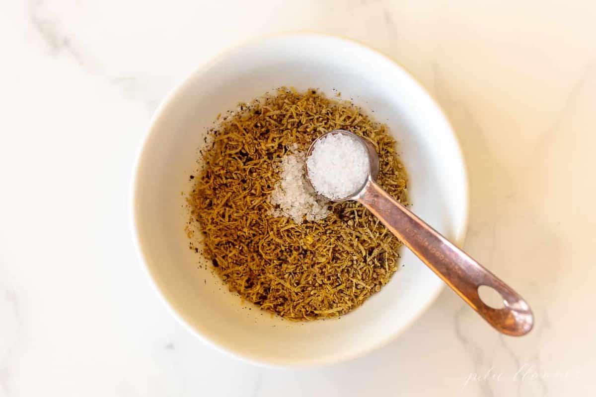 A white bowl with a teaspoon to the side, bowl filled with lemon pepper seasoning.