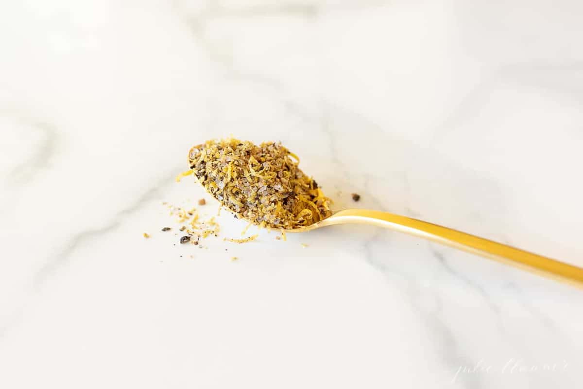 A gold spoon on a marble surface, filled with fresh lemon pepper seasoning.