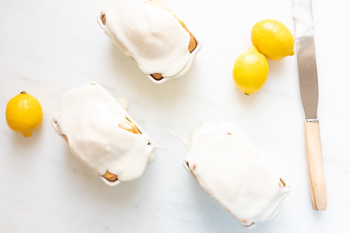 Lemon-glazed loaves of bread with whole lemons and a knife on a white surface, ready to share their lemon glaze icing recipe.