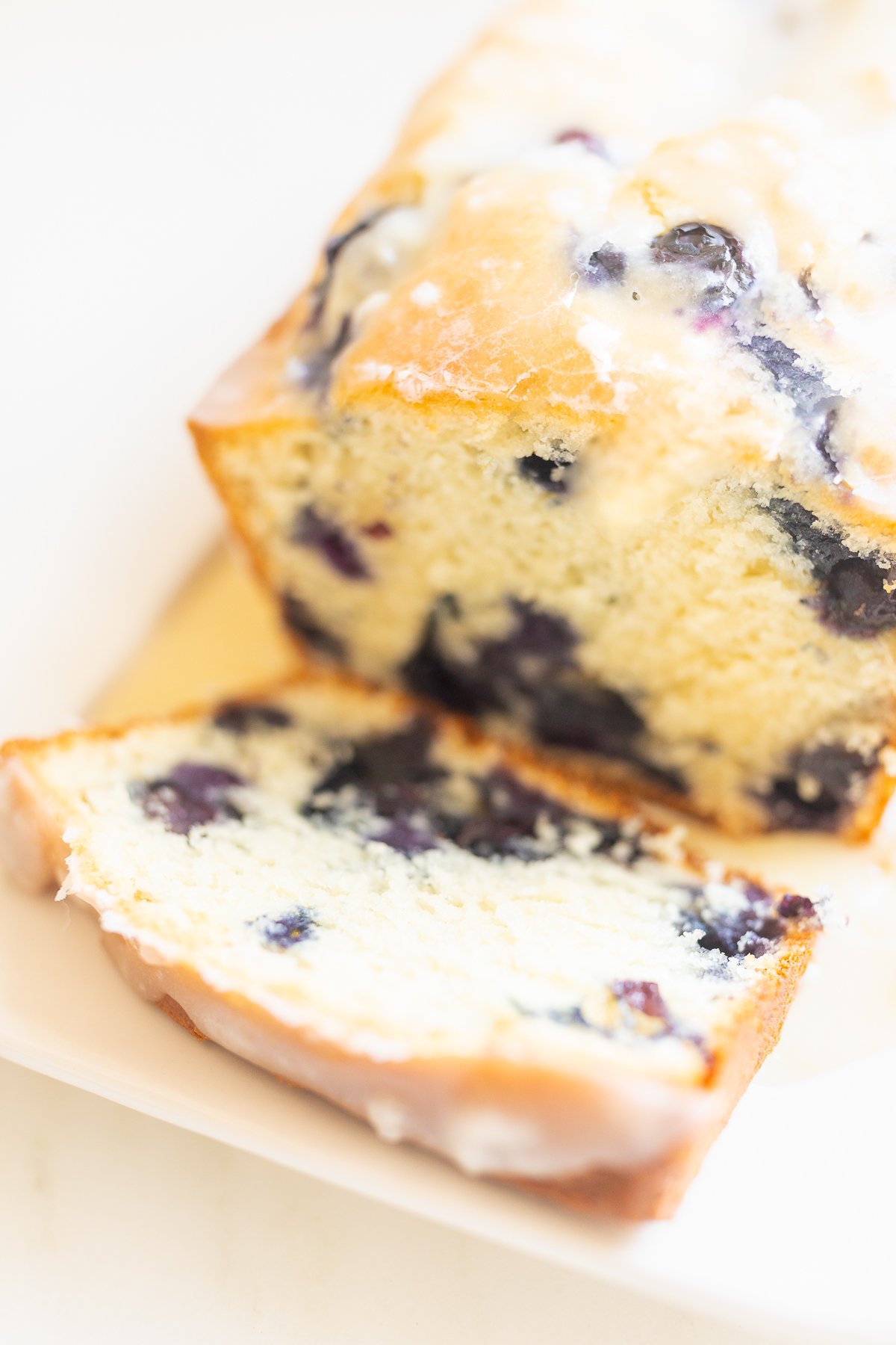 A close-up of a sliced blueberry loaf cake with lemon glaze on a plate.