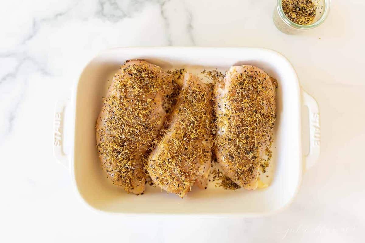 Three raw chicken breasts covered in lemon pepper seasoning in a white baking dish, white basting brush on the top.