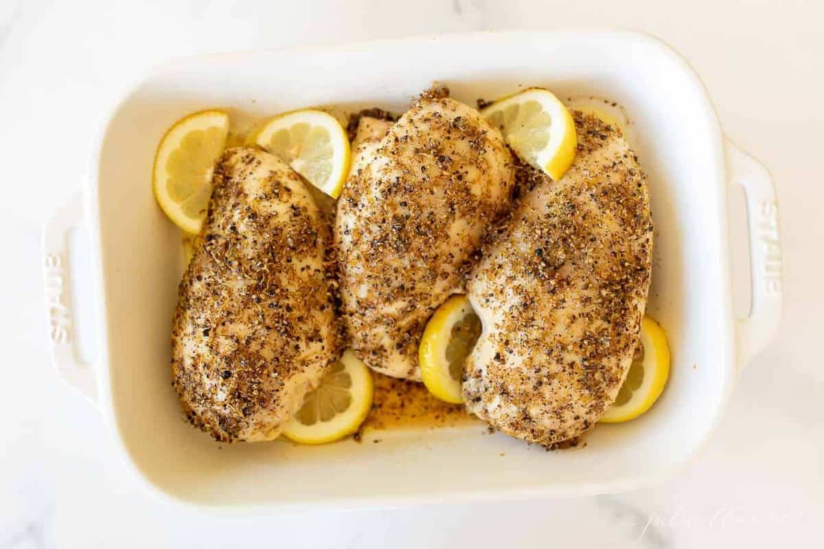 A white dish filled with lemon pepper chicken marinade on chicken breasts, slices of lemon alongside.